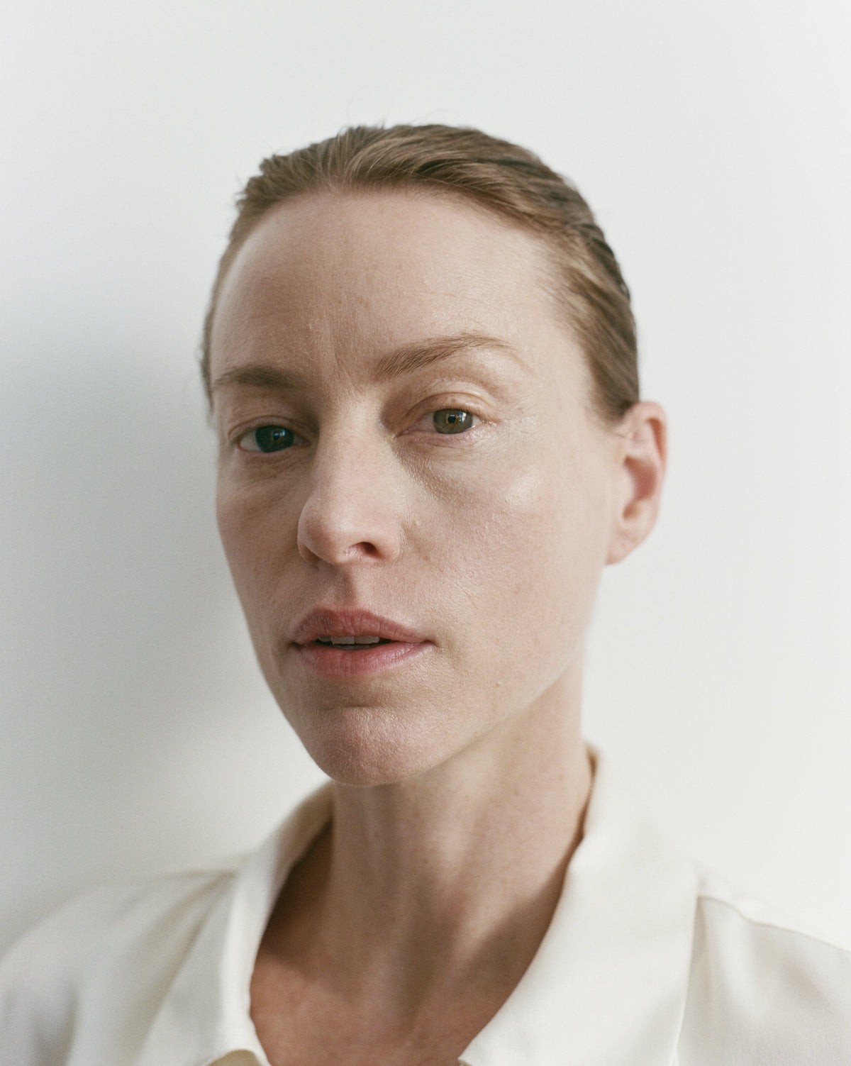 A close-up portrait of a woman with her hair pulled back, wearing a white collared shirt against a plain light background.