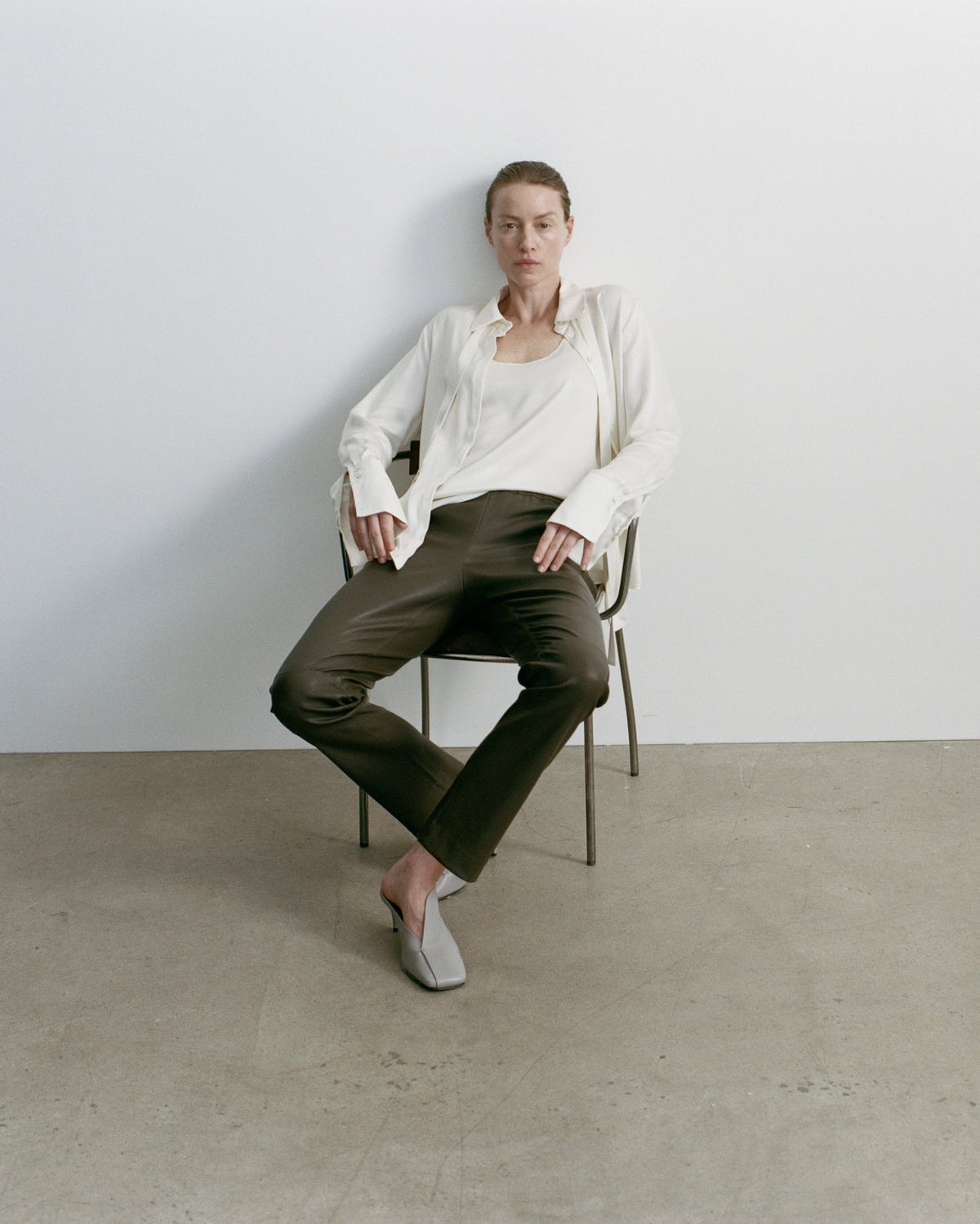 A model sits on a minimalist metal chair against a white wall and concrete floor, wearing a white long-sleeve shirt, dark brown trousers, and white slip-on shoes.